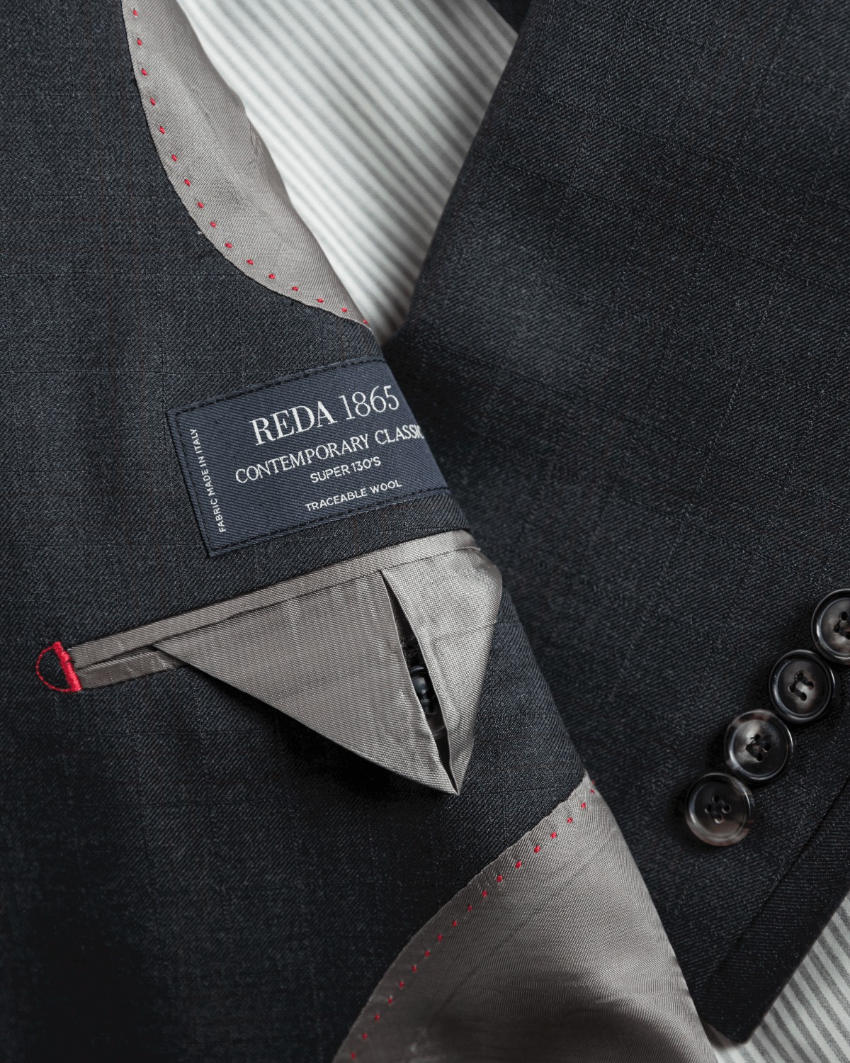 西裝內襯胸袋局部特寫，展示黑色織標印有 REDA 1865 CONTEMPORARY CLASSIC Super 130's TRACEABLE WOOL 字樣，周邊點綴著密麻的紅色手針點腳縫線，灰色裡布折疊出挺拔的三角袋蓋
